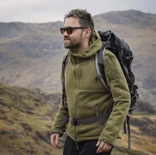 Person with beard wearing green hoodie and sunglasses with backpack