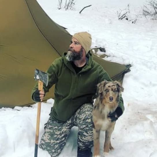 Person with beard wearing green hoodie and beanie in snowy environment with tent and dog
