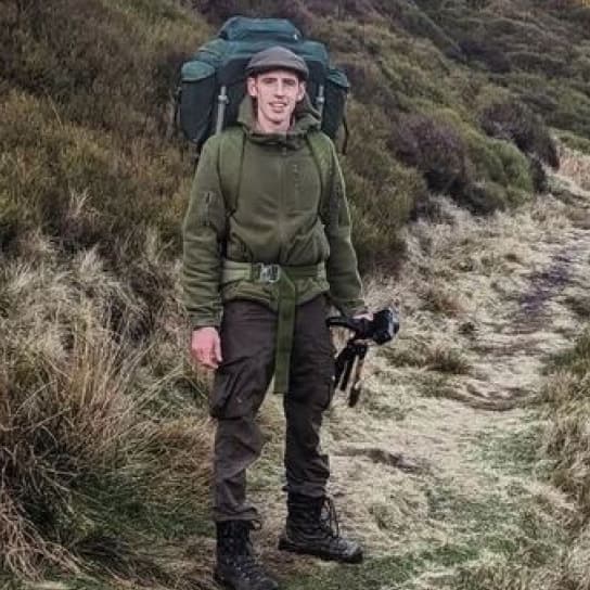 Person with beard wearing green hoodie and backpack on hillside